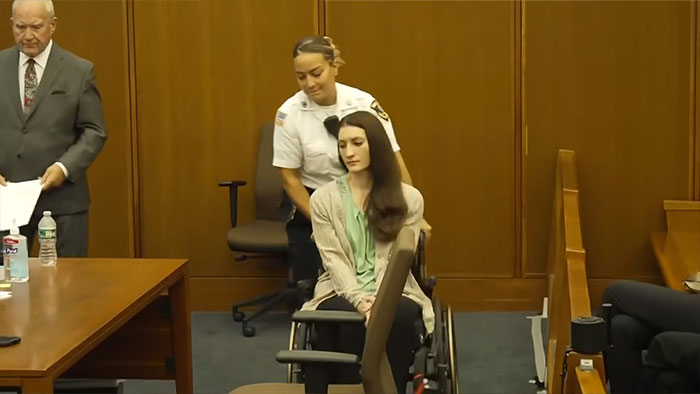 Lindsay Clancy is pushed in a wheelchair by a female officer in the courtroom, with another man walking nearby. Lindsay Clancy is pushed in a wheelchair by a female officer in the courtroom, with another man walking nearby.