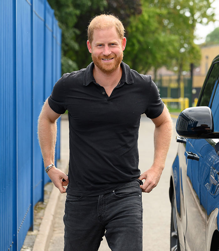 Prince Harry in a black polo and jeans, smiling, after addressing his kids' schooling situation.