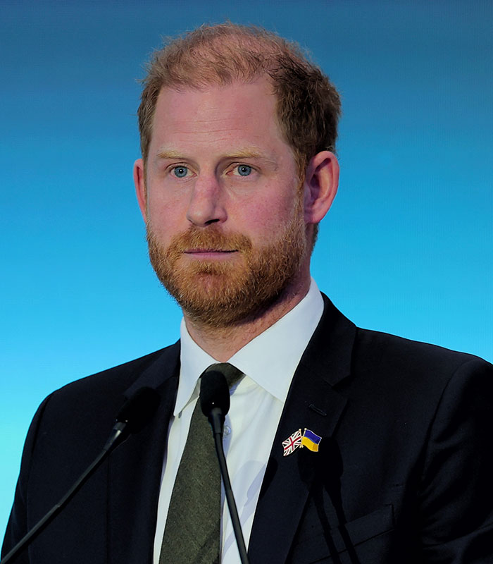 Prince Harry at a podium, looking serious, part of the Meghan and Harry story.