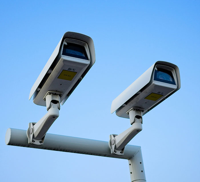 Two white security cameras on a pole against a clear blue sky, overlooking a public area.