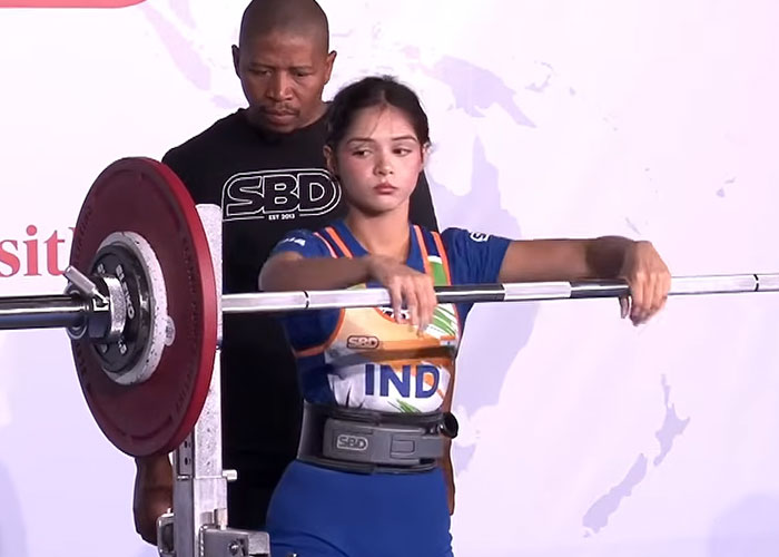 A female weightlifting medalist in a blue uniform, wearing a lifting belt, performs a squat with a barbell on her shoulders. A female weightlifting medalist in a blue uniform, wearing a lifting belt, performs a squat with a barbell on her shoulders.