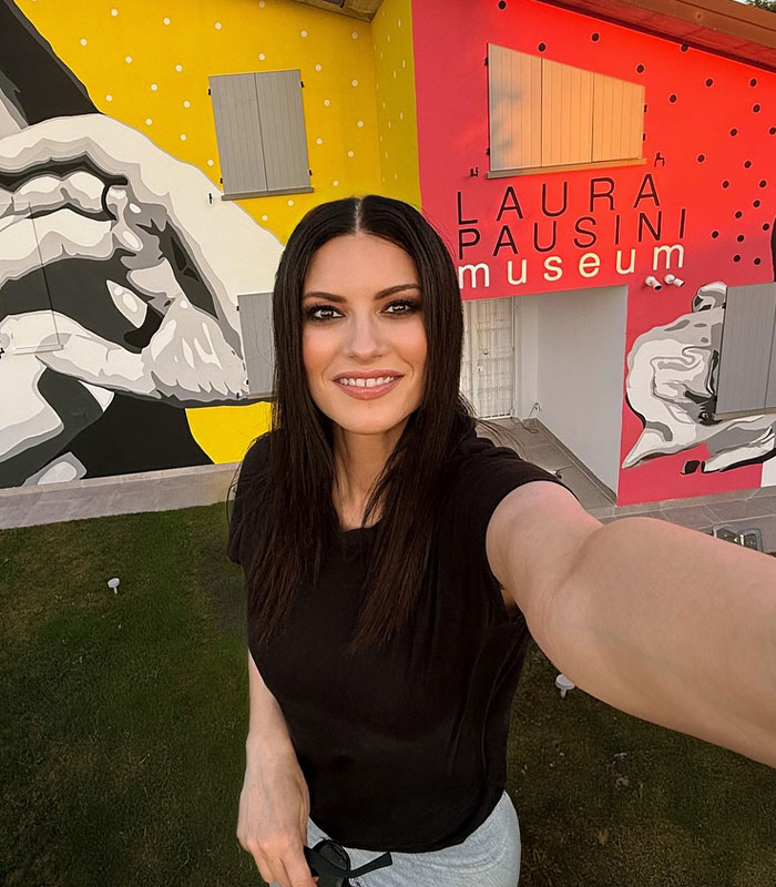 Laura Pausini smiling in front of a colorful mural, capturing the essence of Instagram versus reality.