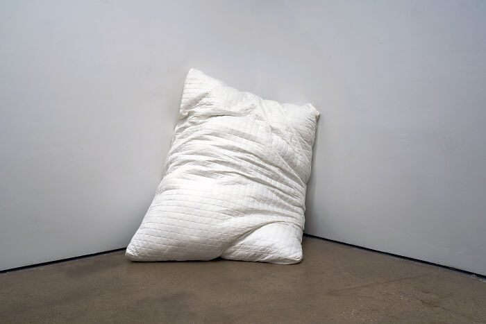 A large white pillow leaning against a corner of a gallery room as part of unusual sculptures
