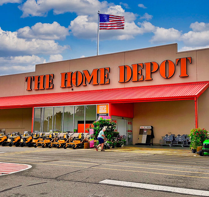 A worker walks towards The Home Depot, a large home improvement store under a blue sky with an American flag flying.