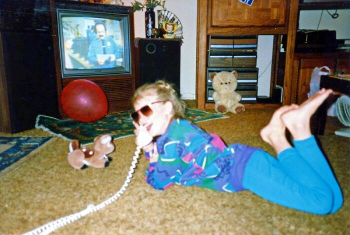 A girl in sunglasses lies on a carpet talking on a retro tech phone, with a TV in the background, capturing the 80s weirdness.