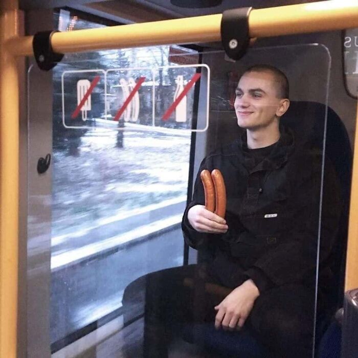 A man with a shaved head smiles, holding two hot dogs on a bus, a hilarious photo for the Internet.