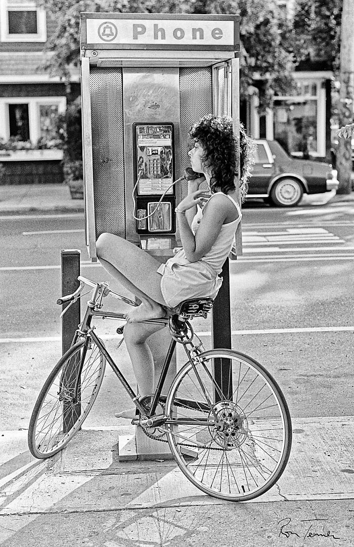 A woman sitting on her bicycle next to a public phone booth, a common sight for 80s retro tech communication.
