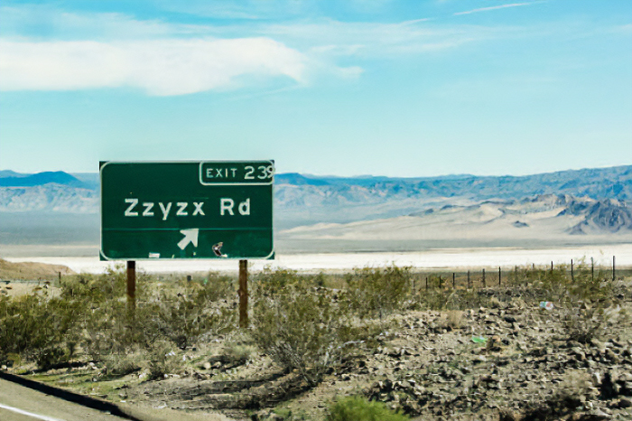 A green road sign for Zzyzx Rd under a blue sky, a chaotic sign spotted in the wild.