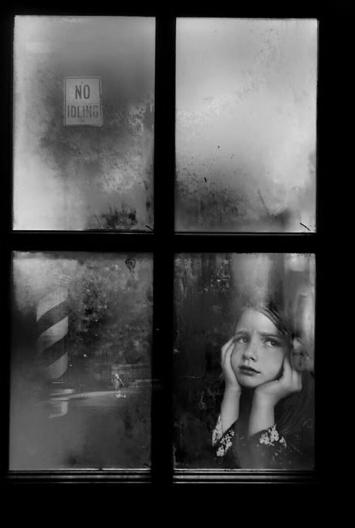 A young girl looks out a grimy window, resting her chin on her hands, a visual story from Taylor Coble's street photos.