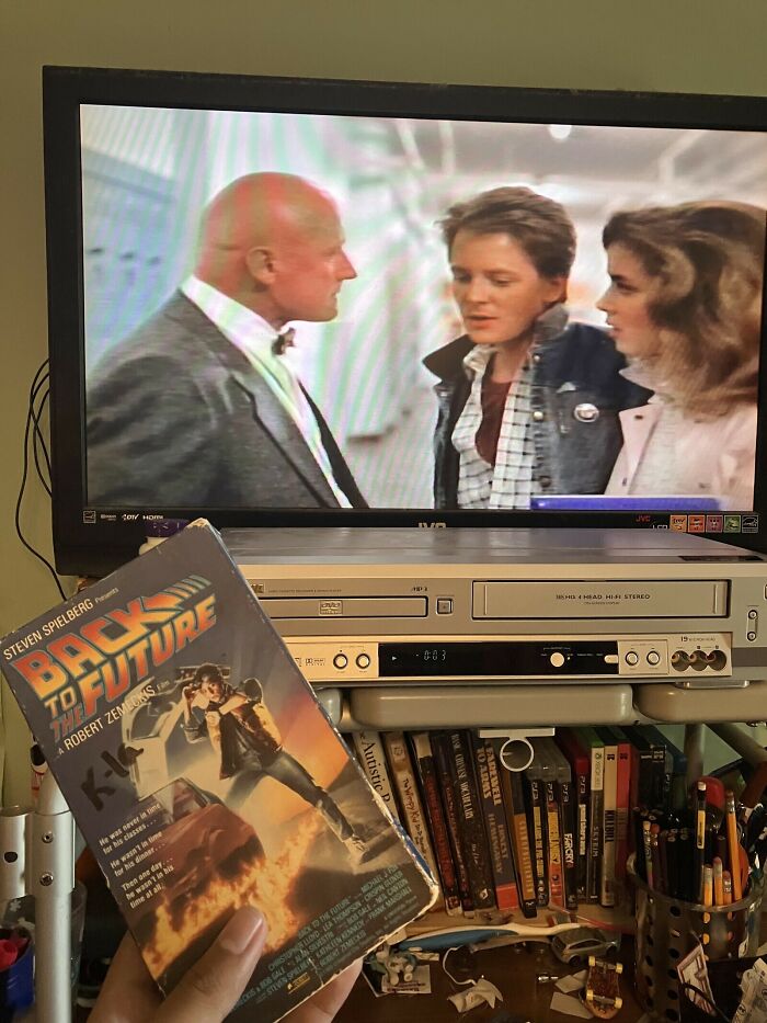 A hand holding a VHS tape of Back to the Future in front of a TV playing the movie, showcasing 80s retro tech.