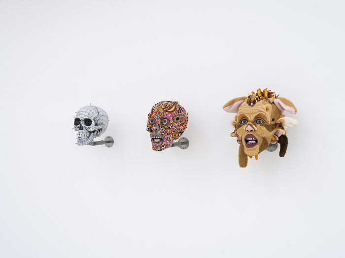 Three unusual sculptures displayed on a wall starting from a white skull to a detailed plush face