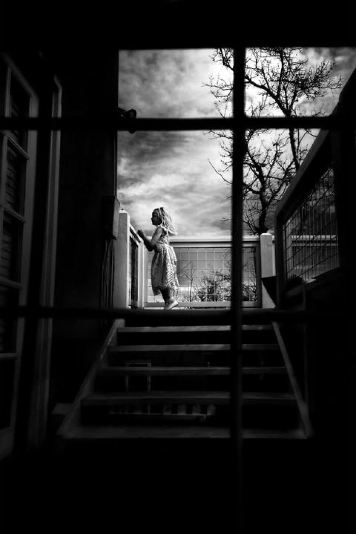 A girl stands on a porch with steps, silhouetted against a cloudy sky, a compelling visual story in street photos.