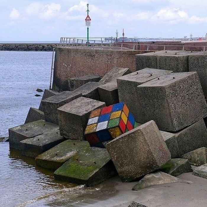 An artistic street art display of a giant Rubik's cube among concrete blocks near the ocean.