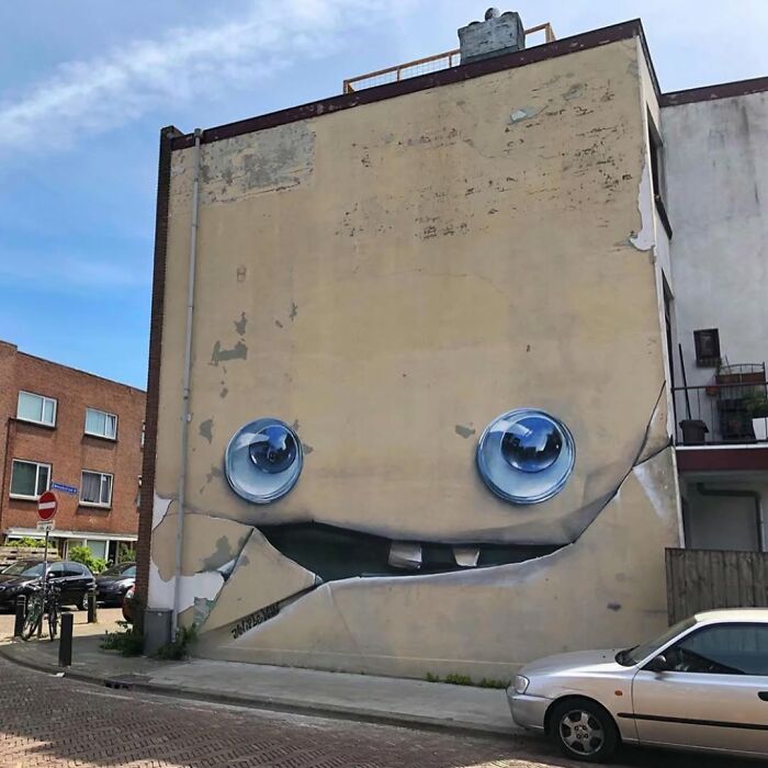 A large-scale street art mural on the side of a building, depicting a whimsical face with big blue eyes and a playful mouth.