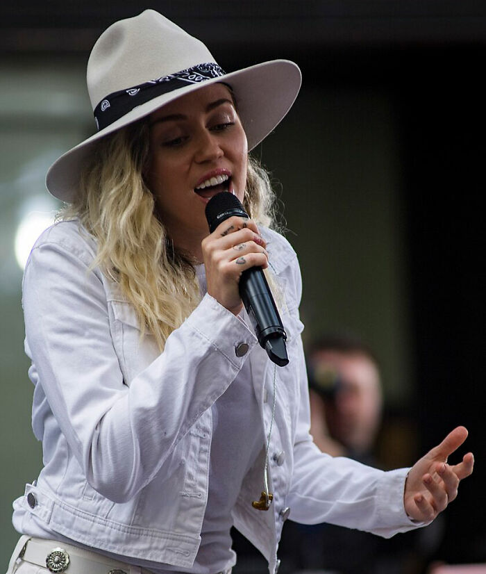 Miley Cyrus singing into a microphone, wearing a white hat and jacket, perfect for celebrity hot takes content.