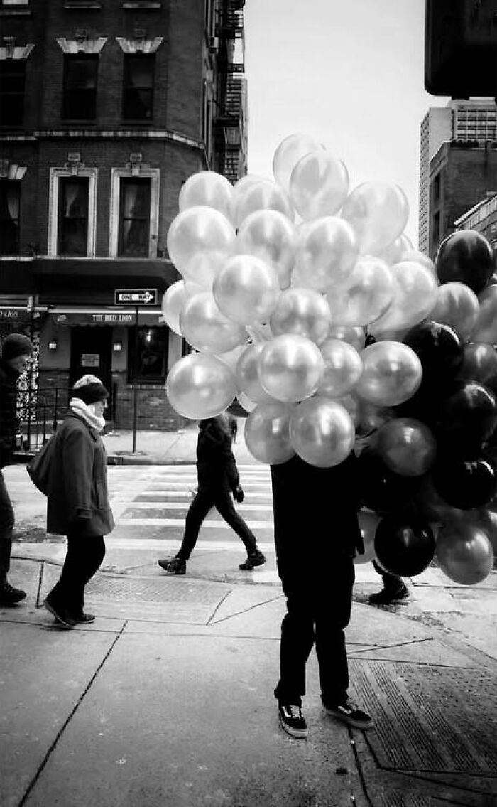 Black and white street photo capturing a person carrying many balloons on a city street, by Taylor Coble.