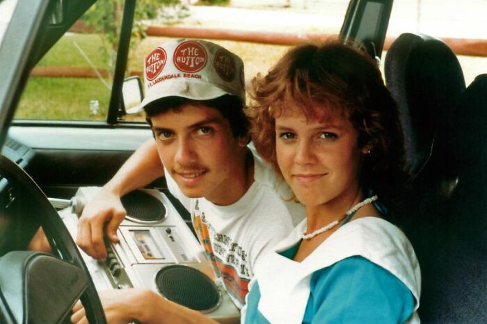 A couple sits in a car, holding a boombox, showcasing '80s retro tech and style.