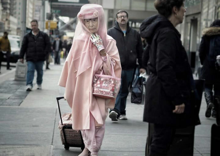 Taylor Coble street photo featuring an elderly woman in a pink outfit pulling a small suitcase on a city street.