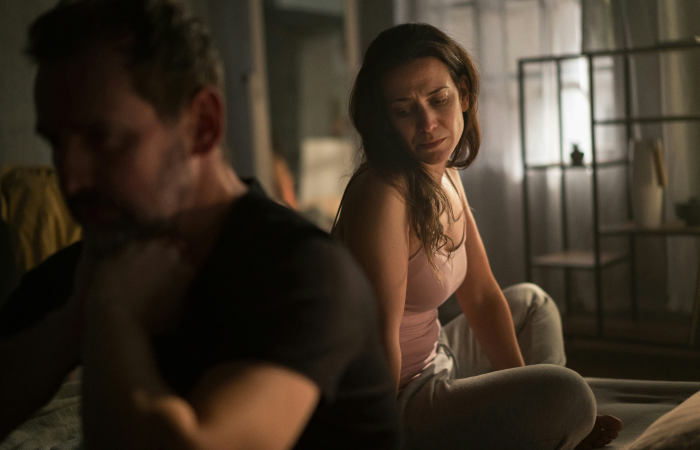A wife looks distressed, sitting behind her husband who refuses to listen, highlighting their marital conflict regarding his son. A wife looks distressed, sitting behind her husband who refuses to listen, highlighting their marital conflict regarding his son.