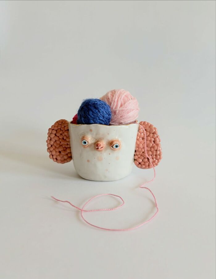 Cute ceramic sculpture cup with pink ears holding balls of yarn, ideal as functional and weird home decor.