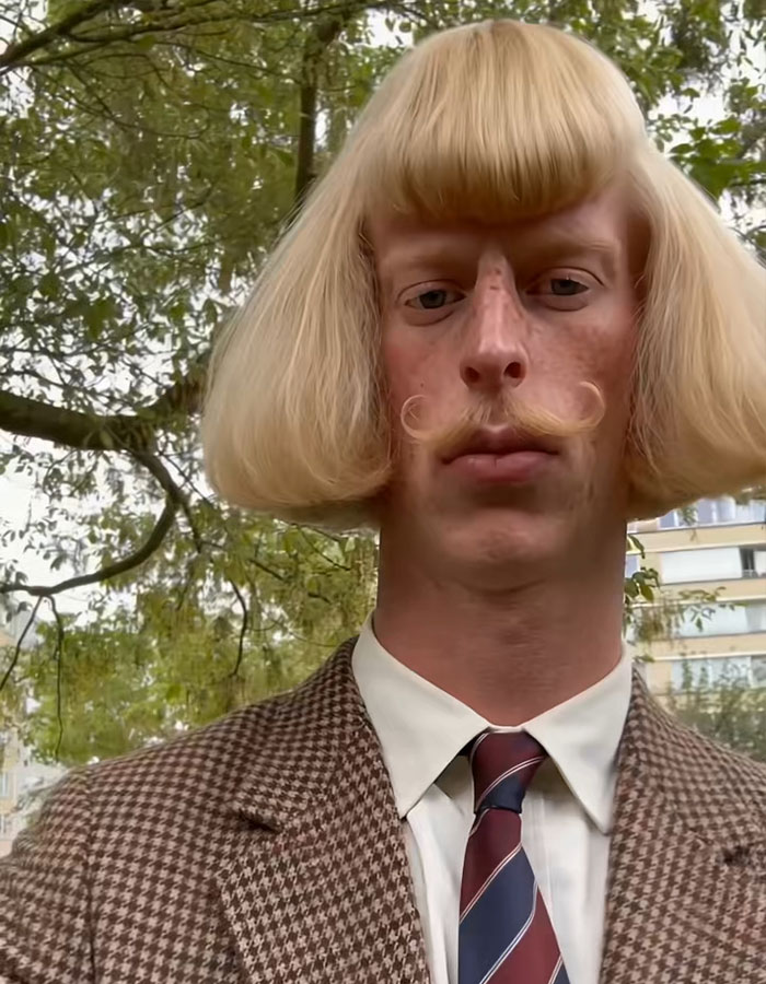 A mysterious influencer with a blond pageboy haircut and mustache looks directly at the camera outdoors