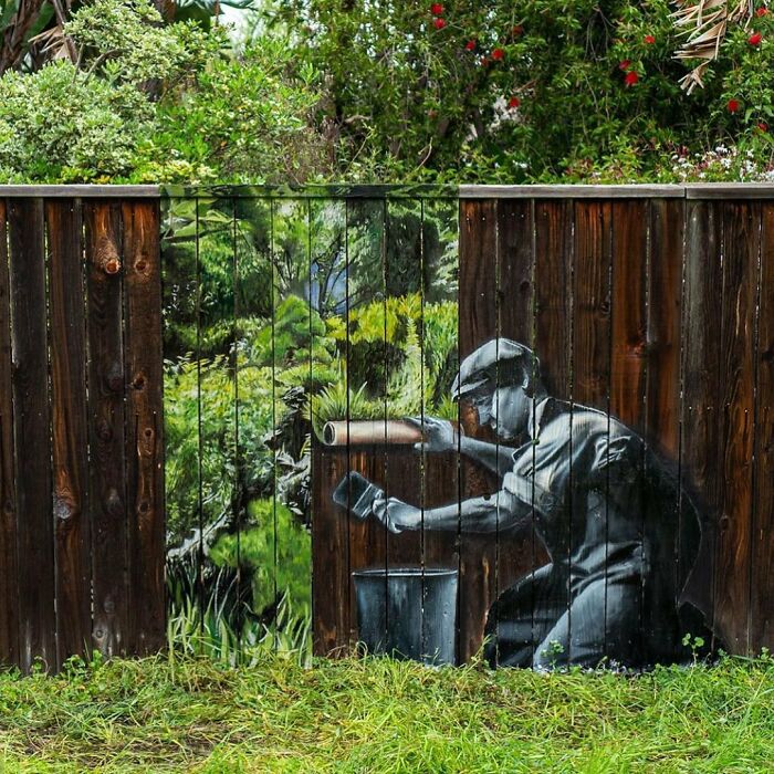 A creative street art piece on a wooden fence shows a painter adding a vibrant green landscape to a natural setting.
