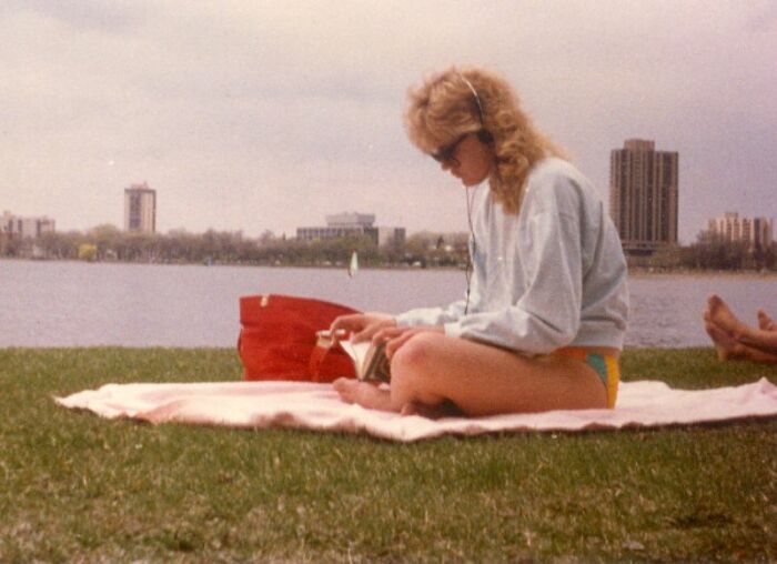 A woman in a swimsuit and sweatshirt sits by a lake, reading and listening to music on retro tech headphones from the '80s.