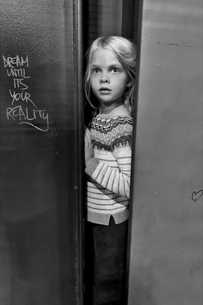 Black and white street photo by Taylor Coble featuring a young girl peering from behind a wall with graffiti.