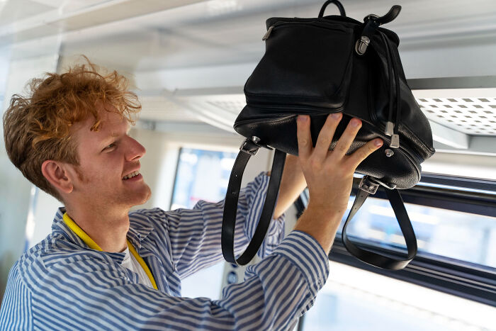 A young man placing his black backpack in the overhead bin on a flight next to his plane seatmate.