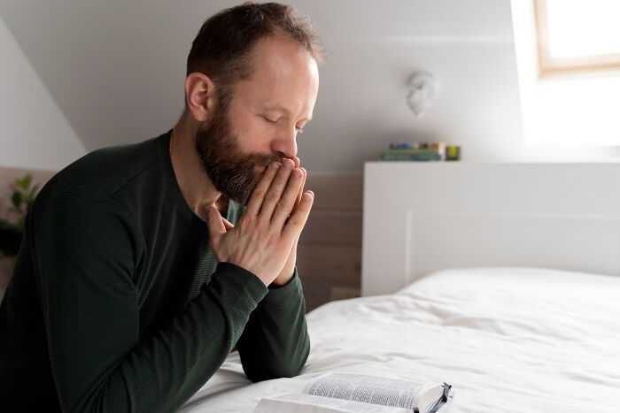 A bearded man with closed eyes prays or meditates by an open book on a bed, showing an unexpected spiritual life story.