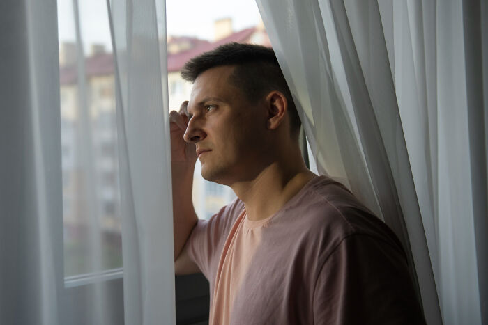 A thoughtful man looking out of a window behind sheer curtains, contemplating ridiculous arguments.