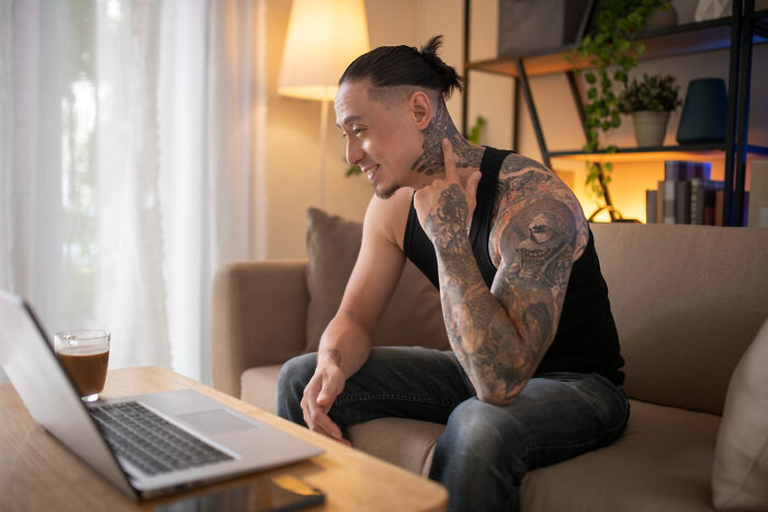 A tattooed man smiling while having a video call on his laptop, discussing ridiculous arguments.