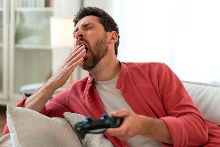 A tired man yawning while playing video games, showing one of the dumb ways people ruin their lives.