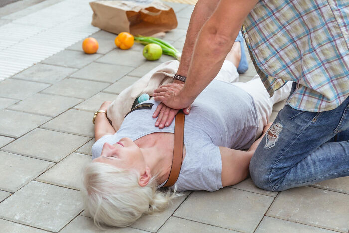 A man performs CPR, a standard medical procedure, on a woman lying on the pavement, with groceries spilled nearby.
