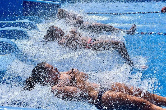 A street photographer captures three people in a swimming pool, covered in splashing water, enjoying a fun day.