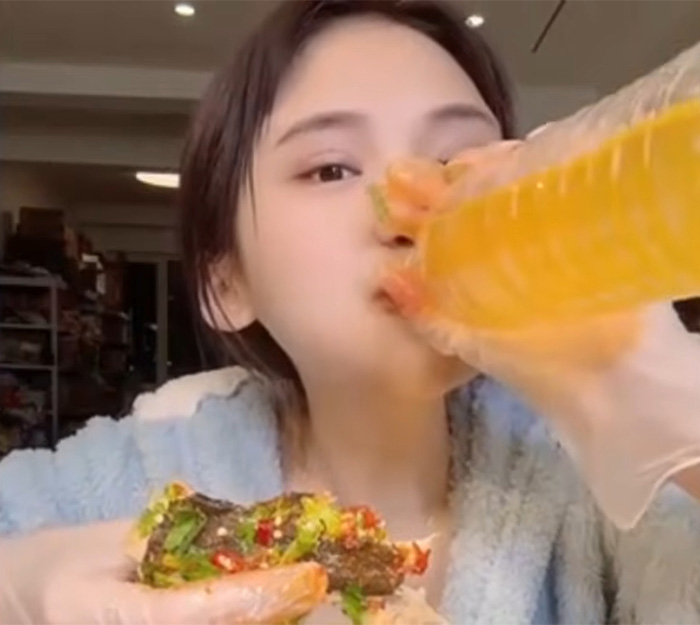 A young mukbang star eats a large, spicy burger and drinks from a bottle, showing extreme eating.