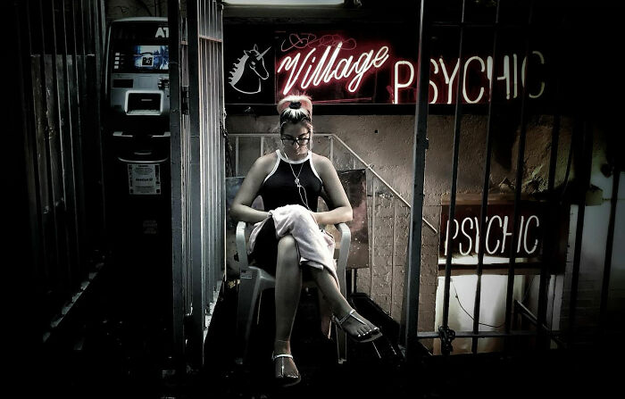 A neon-lit street photo by Taylor Coble showing a woman sitting in a chair outside a Village Psychic business.