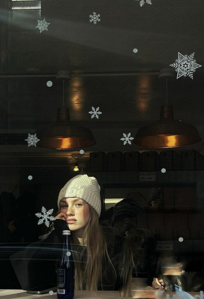 Taylor Coble street photo of a young woman in a white hat looking out a window adorned with snowflake decals.