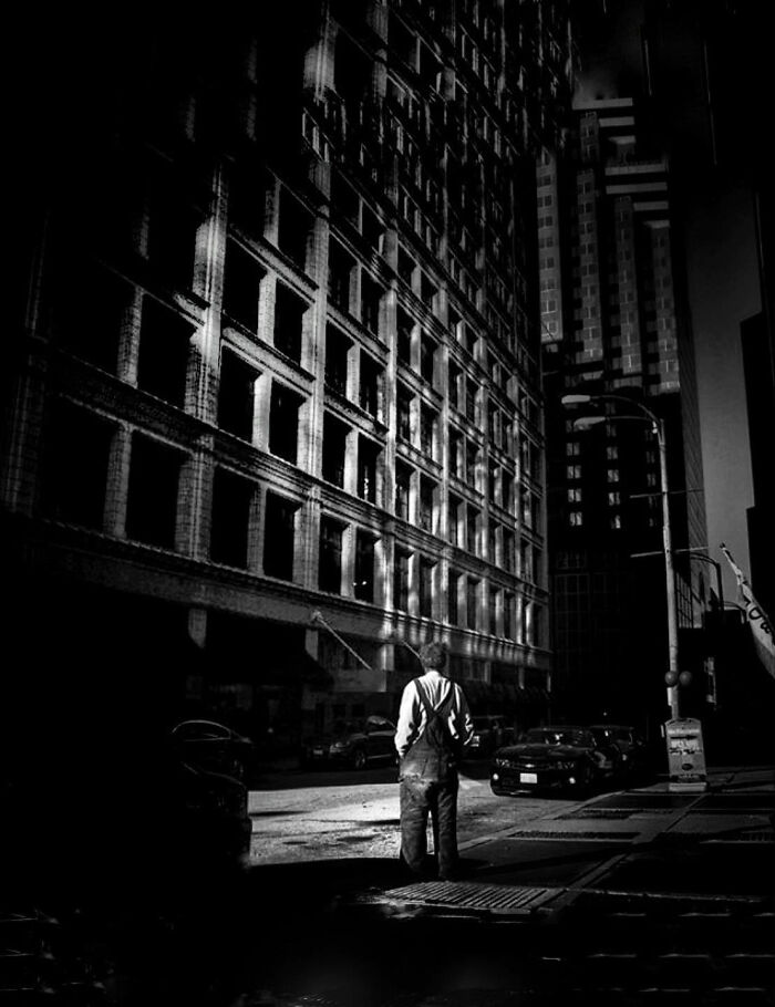 A lone figure stands on a dark street, surrounded by tall city buildings, a poignant street photo capturing a visual story.