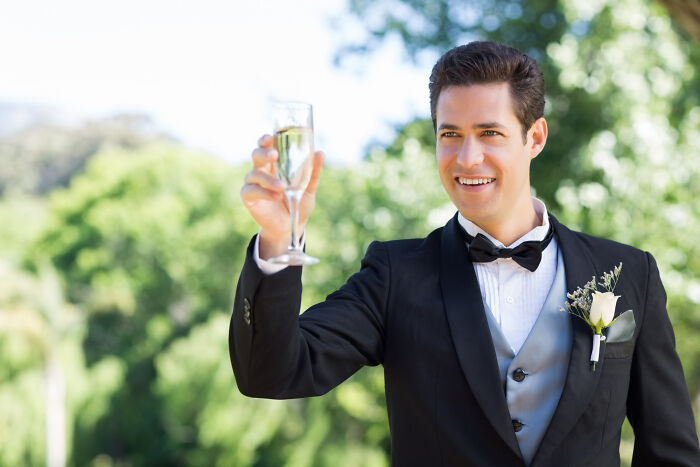 A groom in a black tuxedo raises a champagne glass for a toast, celebrating joy at weddings.