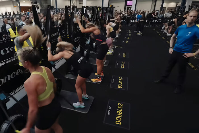 Athletes intensely working out on machines during a HYROX race, watched by spectators and staff.