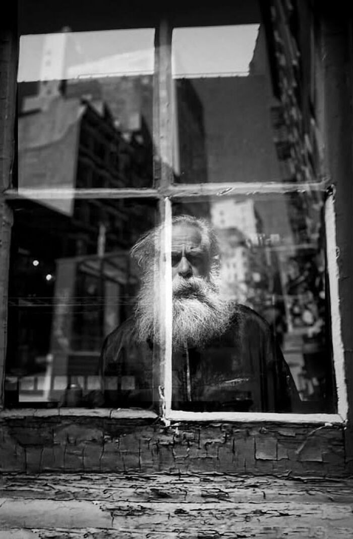 A man with a long white beard looks out a weathered window in a black and white street photo by Taylor Coble.