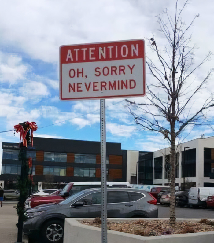 A funny sign saying ATTENTION OH, SORRY NEVERMIND is posted next to a bare tree in a parking lot.