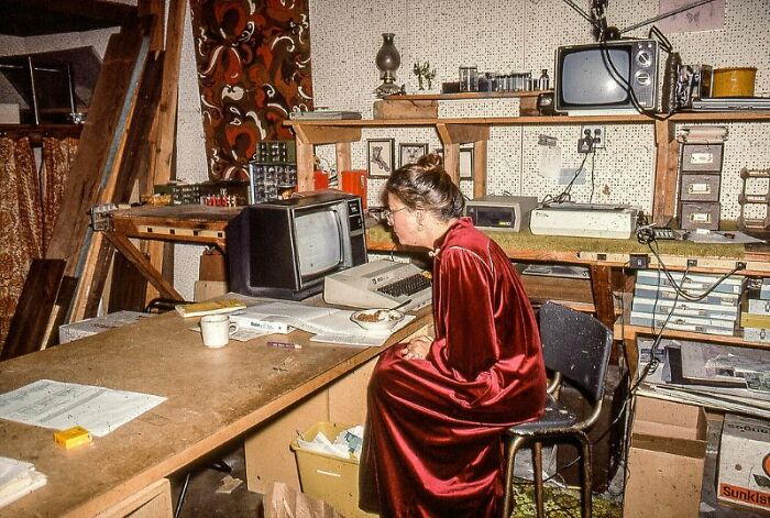 A woman in a red robe sits at a desk with an old computer and CRT monitor, surrounded by 80s retro tech.