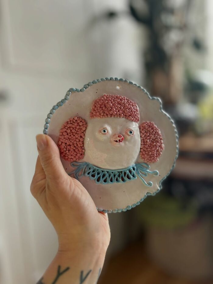 A hand holding a circular pink poodle wall plate, displaying weird ceramic sculptures for home decor.