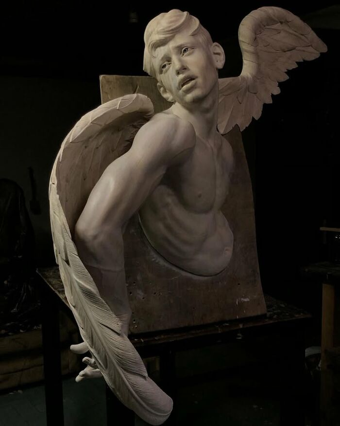 A lifelike wood sculpture of a muscular winged man leaning forward, created by a Polish artist