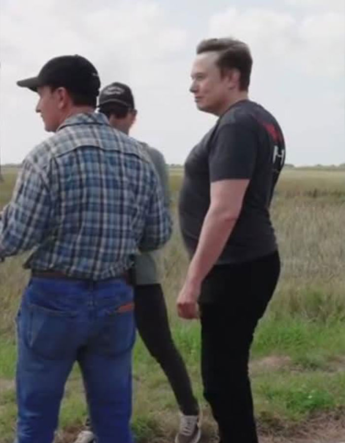 Elon Musk standing outside with colleagues, side view showing posture