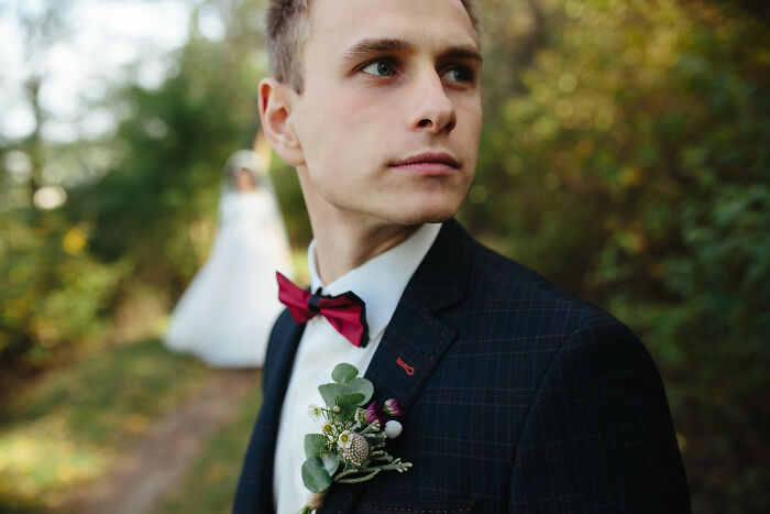 A close-up of a groom wearing a bowtie and boutonniere with a bride blurred in the background at weddings