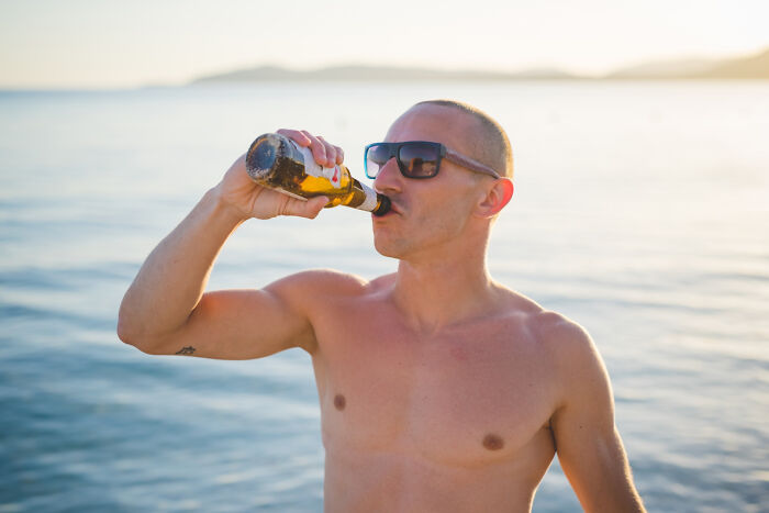 A man wearing sunglasses drinks a beer by the ocean, illustrating an unexpected life story unfolding by the water.