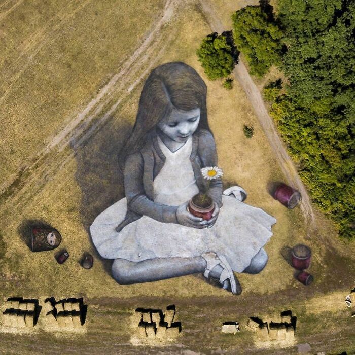 Large scale street art of a young girl in a field, holding a potted flower, viewed from above.
